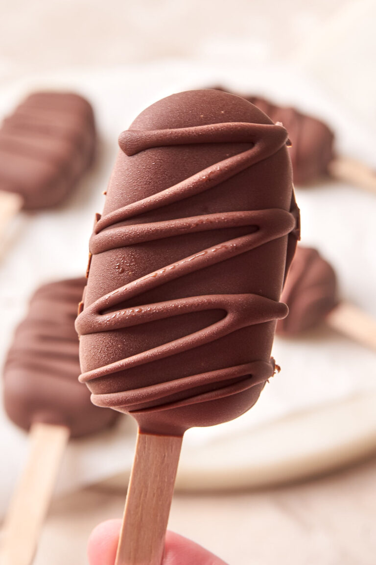 A hand holding a chocolate ice cream bar on a stick, decorated with a drizzle of chocolate.